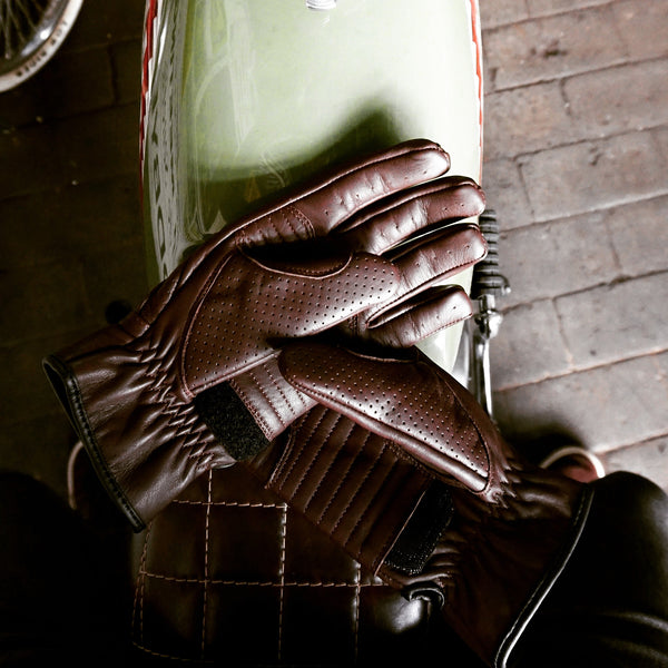 A pair of Speed motorcycle gloves by 78 Motor Co., featuring a mix of brown leather with black accents and a velcro strap on the wrist sitting top of a vintage motorcycle.