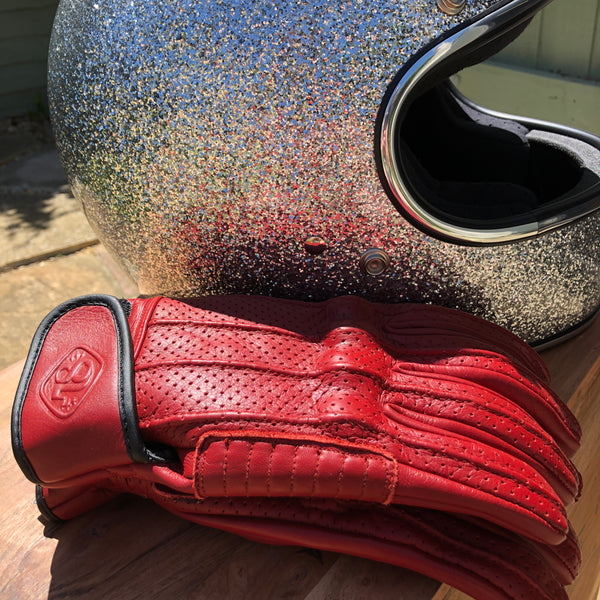 A pair of Speed motorcycle gloves by 78 Motor Co., made of red perforated leather with knuckle protection and a velcro strap on the wrist with the number '78' embossed on the strap with a sparkly motorcycle helmet.